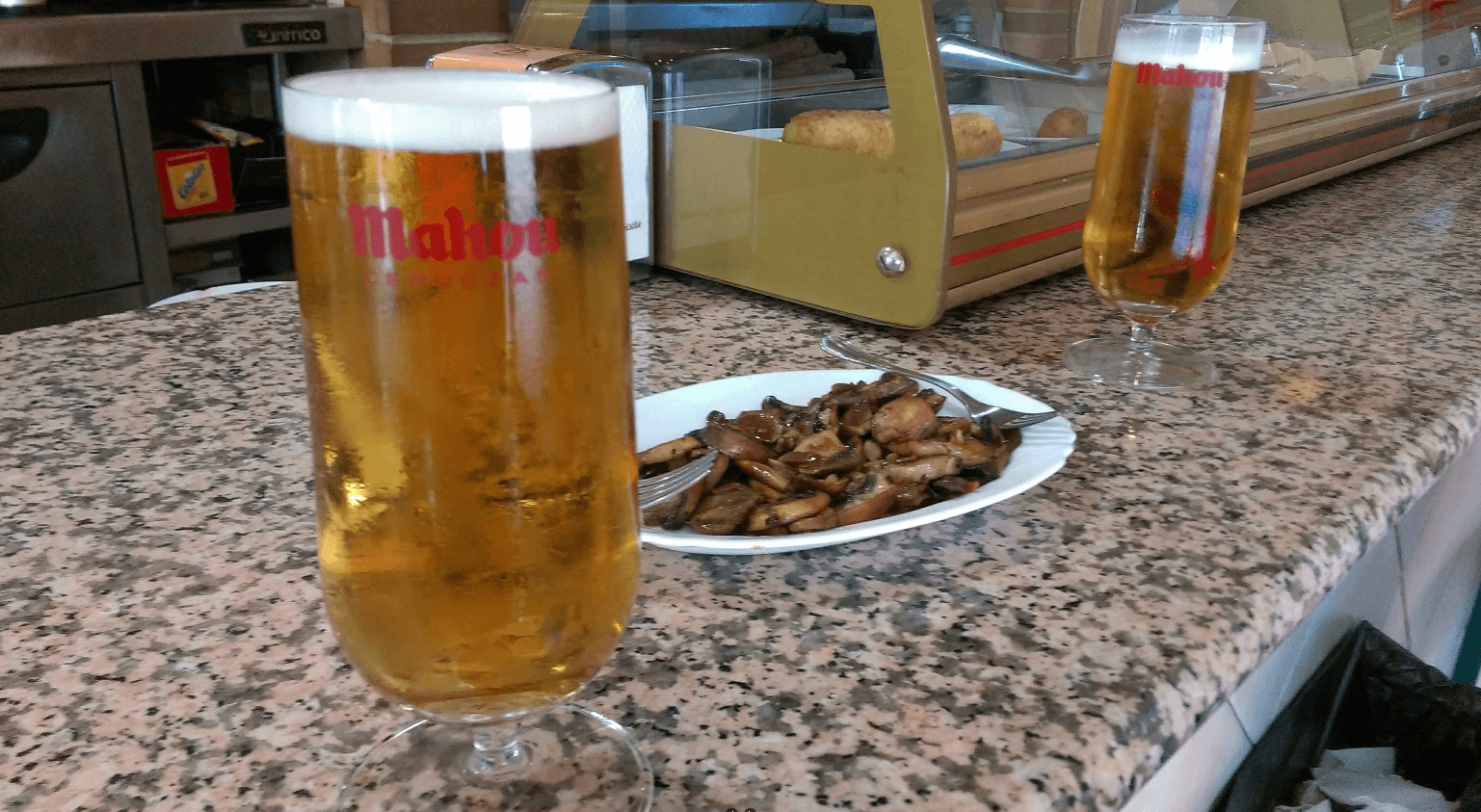 Dos cañas de cerveza Mahou junto a una tapa de champiñones sobre barra de granito.