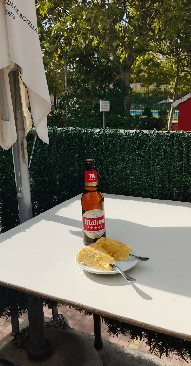 Botella de cerveza Mahou y tortilla de patatas en una mesa blanca de terraza soleada.