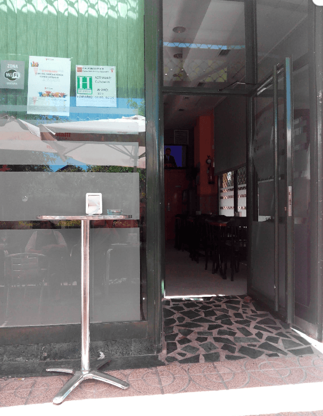 Entrada de una cafetería con mesa alta metálica y carteles informativos en el cristal.